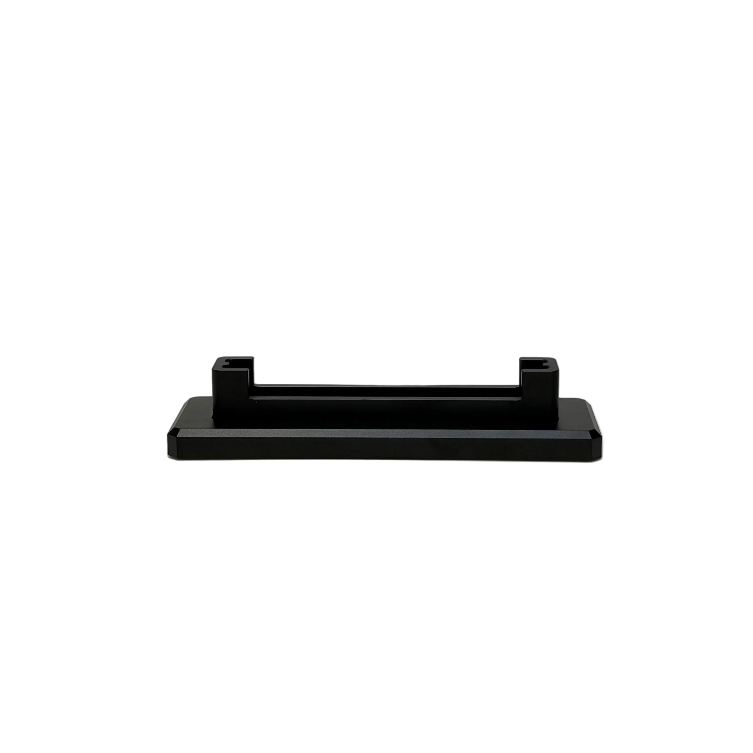 A small black plastic rectangular mounting bracket or clip, shown from the side, resting on a round white display platform against a textured light beige wall.