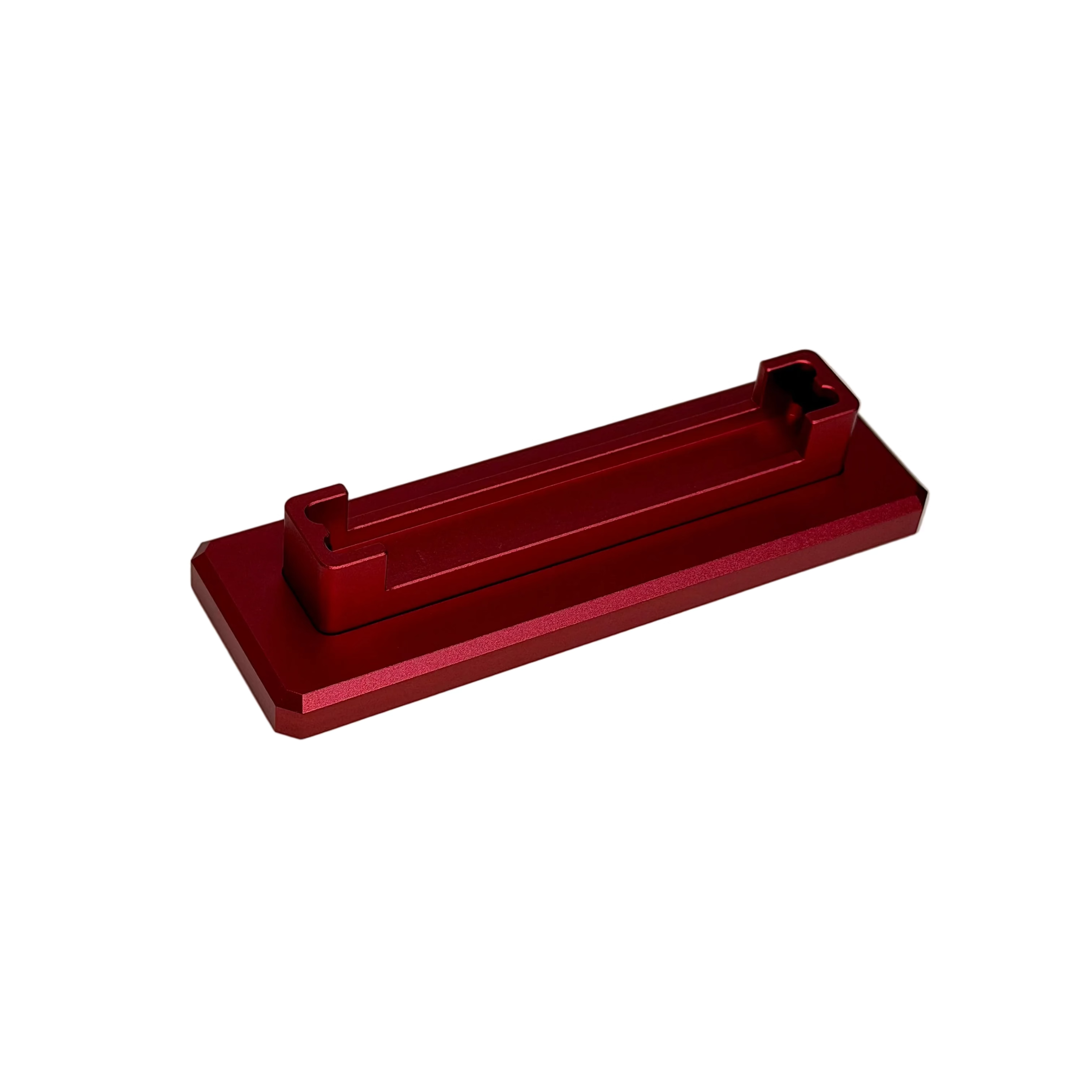A small red anodized metal bracket or clip with a rectangular base and two small upright tabs, photographed on a round off-white display surface against a white background.